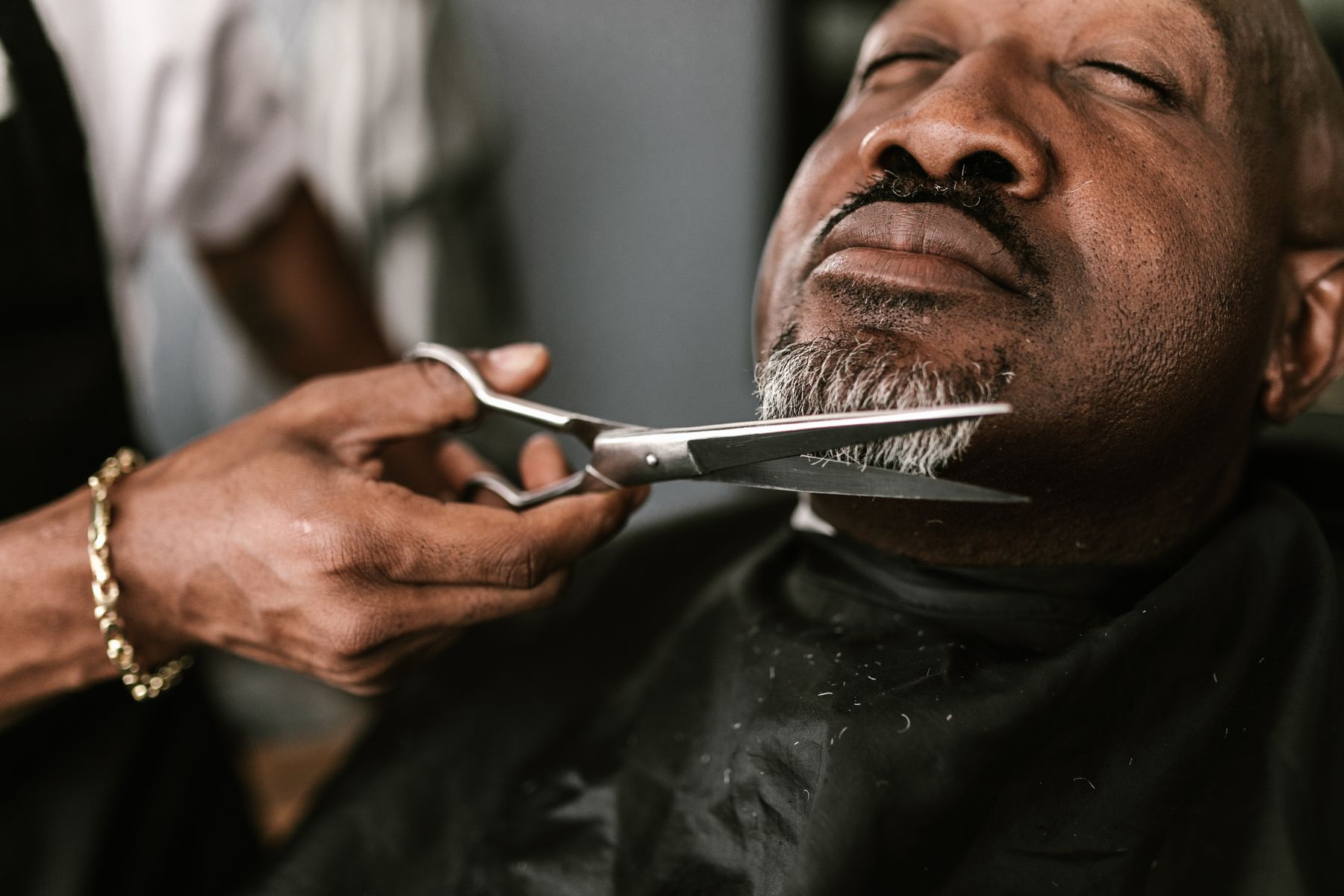 A barber trimming a client's grey beard with scissors