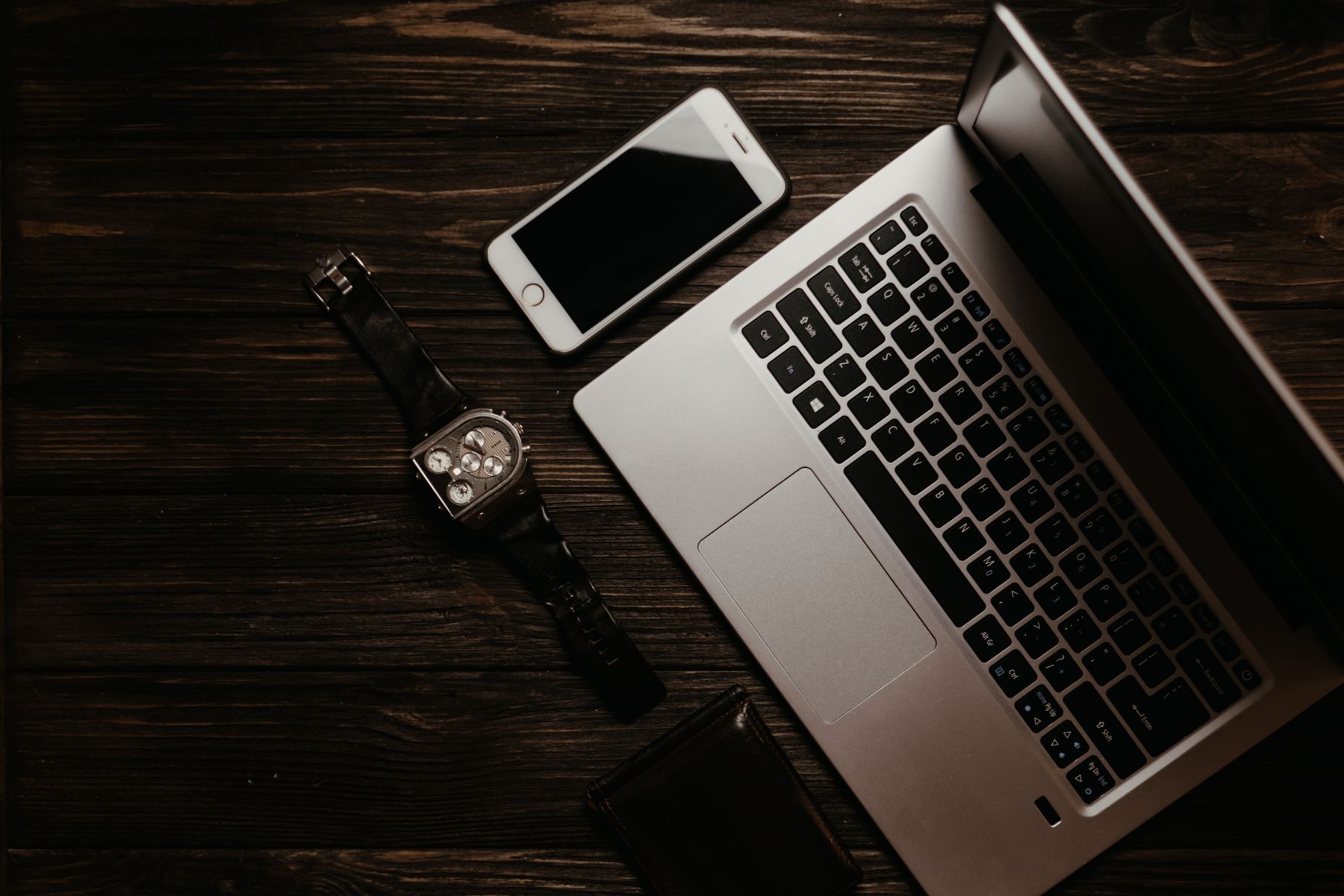 A laptop, phone and watch on dark wood